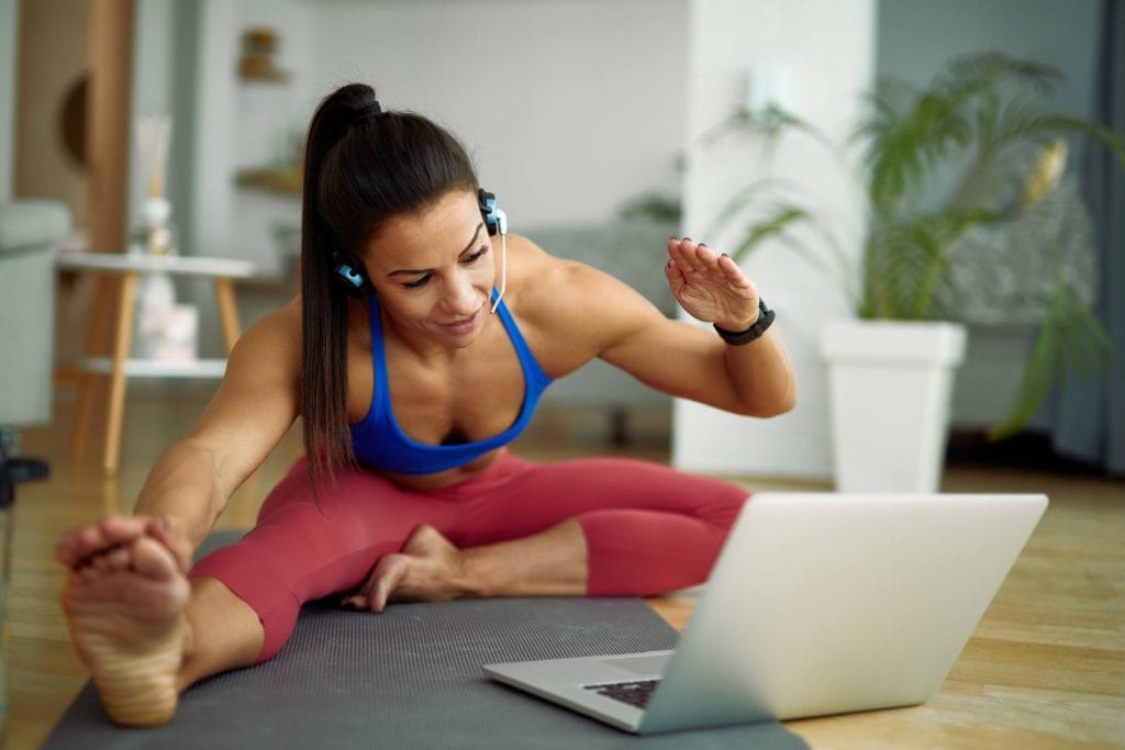 Woman on a Pilates mat at home stretching and waving to her instructor during a live online Pilates class on her laptop